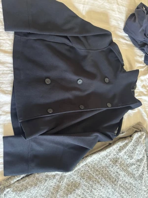 Svart dubbelknäppt rock från Gina Tricot - Stilren svart rock från Gina Tricot med dubbelknäppning och stora knappar framtill. Kortare modell med klassisk krage och raka ärmar. Perfekt för dig som vill ha en enkel men snygg look. Materialet är tjockt och känns slitstarkt.