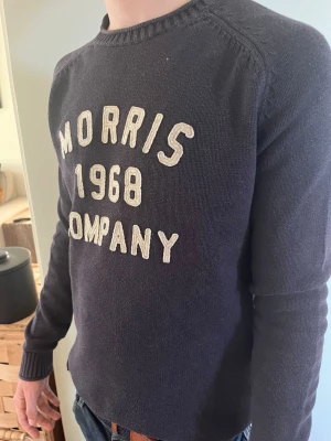 Mörkblå stickad tröja Morris - Mörkblå stickad tröja från Morris med vita broderade bokstäver på bröstet där det står 'Morris 1968 Company'. Tröjan har rund halsringning, långa ärmar och ribbade muddar. Perfekt för en avslappnad och stilren look.        Skickas inom 1-3 dagar vid beställning 