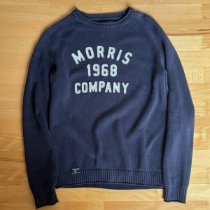 Morris mörkblå tröja med tryck - Mörkblå stickad tröja från Morris med vit text 'Morris 1968 Company' på bröstet. Tröjan har rund halsringning, långa ärmar och ribbade muddar vid ärmslut och nederkant. Sparsamt använd!