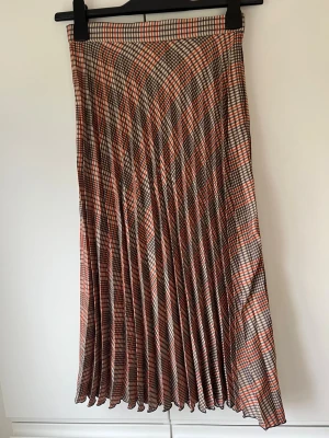 Plisserad rutig kjol i beige och rött - Säljer en plisserad kjol med rutigt mönster i beige, rött och blått. Kjolen har hög midja och ett klassiskt, tidlöst uttryck som passar perfekt till både stickade tröjor och t-shirts. Sold dragkedja i midjan. 