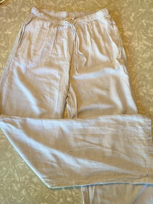 Randiga pyjamasbyxor - Ljusblå och vita pyjamasbyxor med tunna vertikala ränder och dragsko i midjan. Byxorna har avslappnad passform och är tillverkade i ett mjukt, lätt tyg som känns skönt mot huden. Perfekta för mysiga hemmakvällar eller lata morgnar. Har ett litet hål längst ned på benet men inte något som syns vid användning. 
