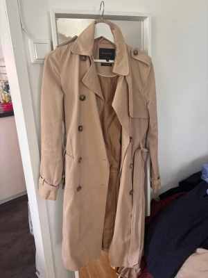 Beige trenchcoat med bälte - Stilren beige trenchcoat med klassisk dubbelknäppning, axelklaffar och bälte i midjan. Jackan har långa ärmar med spännen vid ärmsluten och en snygg krage. Perfekt för dig som vill ha en tidlös och trendig look. Fläckig pris kan diskuteras :)