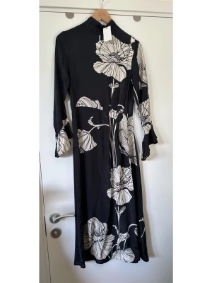 Indiska - helt NY svart klänning med vita blommor - storlek XS - HELT NY underbar svart lång klänning från INDISKA i storlek XS, med ett vackert blommigt mönster i svart och benvitt. Stilren tidlös modell med hög krage och långa ärmar med liten volang vid ärmsluten. Diskret dold dragkedja baktill i nacken och smickrande liten veckning i ena sidan i midjan. Finvävt luftigt tyg med fint fall, i 100% viskos, så bra naturmaterial som andas. Nypris 799:-. Helt ny oanvänd med etikett!