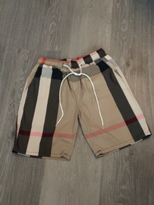 Burberry shorts  - Snygga beige shorts med breda svarta, vita och röda ränder. Shortsen har snörning i midjan och avslappnad passform. Perfekta för sommardagar och har en modern streetwear-vibe.