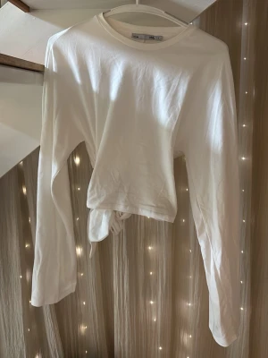 Vit croppad långärmad topp med knyt - Säljer en vit croppad långärmad topp i mjuk bomull. Toppen har rund halsringning och en snygg knytning i midjan som ger en trendig look. Aldrig använd