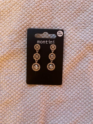 Guldiga glittriga örhängen Montini - Säljer ett par guldiga hängande örhängen från Montini med tre cirkelformade delar på rad. Örhängena är dekorerade med glittriga stenar som ger extra bling och passar perfekt när du vill sticka ut lite extra.