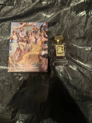 Triumph of Bacchus Eau de Parfum 30ml - Lyxig parfym i en elegant, fyrkantig glasflaska med guldlock och djup röd vätska. Flaskan har en dekorativ etikett med klassiskt motiv och kommer i en konstnärligt designad ask med antikinspirerat tryck. Perfekt för dig som gillar exklusiva dofter och snygg design.