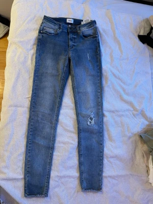 Blå skinny jeans från ONLY - Säljer ett par blå skinny jeans från ONLY med slitningar på ena benet och klassisk femficksdesign. Jeansen har hög midja och är tillverkade i stretchigt denim för en bekväm passform. Perfekta för en trendig och avslappnad look.