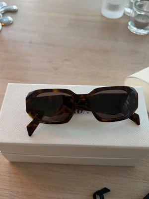 Prada solglasögon - Säljer dessa jättefina solglasögon från Prada. Modellnamn finns på bilderna. De har endast enstaka små repor på bågarna (inga på glaset!) men dessa syns inte vid användning. Äkta! Nypris ca 4900kr.