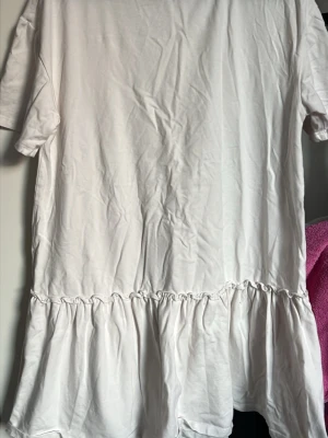 Vit kort klänning med volang - En enkel vit kort klänning i bomull. Klänningen har en volang nedtill som ger en avslappnad och söt look. Perfekt för varma dagar och enkel att styla med accessoarer. Etiketten är borttagen men den är ifrån lager 157 och i storlek M