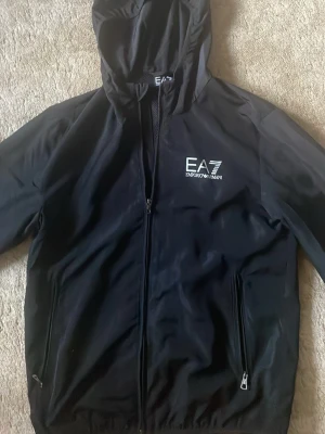 Svart vindjacka från EA7 Emporio Armani - Snygg svart vindjacka från EA7 Emporio Armani med huva och dragkedja framtill. Jackan har meshfoder, två fickor med dragkedja och logga på bröstet. 9/10 skick, knappast använt.