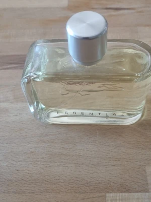 Essential parfym 125 ml - Säljer en stilren parfymflaska med genomskinligt glas och silverfärgad kork. Flaskan rymmer 125 ml och har texten 'ESSENTIAL' på framsidan. Passar dig som gillar klassisk och enkel design. 100 ml kvar 