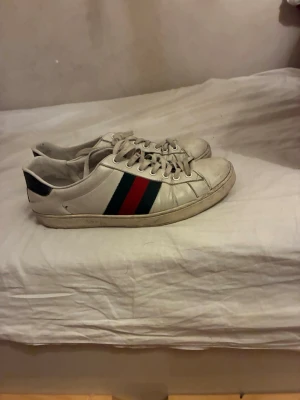 Vita sneakers med röd och grön rand - Snygga vita sneakers i läder med klassiska ränder i rött och grönt på sidan. Skorna har lågt skaft, vita snören och en enkel, stilren design. Perfekta för dig som gillar en clean look med en touch av färg.