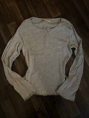 Ljusgrå ribbad långärmad topp - Ljusgrå långärmad topp med ribbad struktur och knappar framtill. Toppen har en lätt vågig kant nertill och vid ärmsluten, samt en rundad halsringning. Perfekt för lager på lager och enkel att styla till olika looks.
