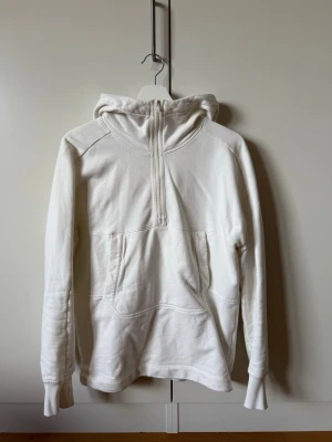 Vit hoodie med glasögon i huvan från C.P Company - Säljer en unik vit hoodie från C.P Company med halv dragkedja framtill och stor känguruficka. Huvan har inbyggda glasögon, vilket ger en cool och futuristisk vibe. Tillverkad i mjukt bomullsmaterial och har långa ärmar med muddar. Den saknar ett snöre som satt på zipen.