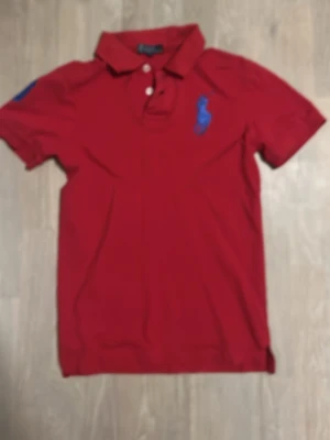 Röd  Ralph Lauren pikétröja med blå detaljer - Klassisk röd pikétröja med blå broderad logga på bröstet och siffran 3 på ärmen. Tröjan har krage, knappar framtill och korta ärmar. Perfekt för en sportig och stilren look.