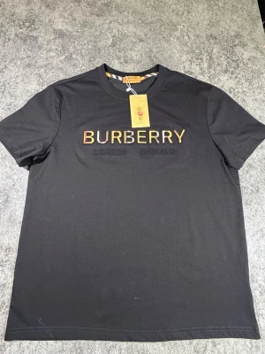 Svart Burberry t-shirt med logga - Snygg svart t-shirt från Burberry med klassisk logga i färgglada bokstäver och texten 'London England' broderat under. T-shirten har rund hals och korta ärmar. Perfekt för dig som vill ha en stilren och exklusiv look.