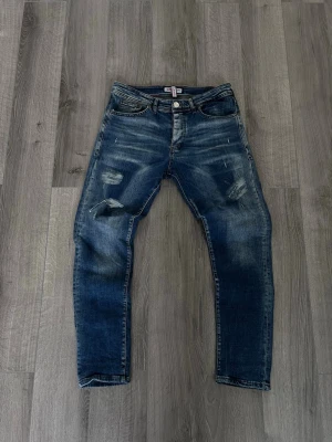 Blåa jenas disqueard - Säljer ett par blå jeans med slitningar och tvättade detaljer. Jeansen har fem fickor, knappgylf och raka ben. Materialet är klassisk denim som ger en cool och avslappnad look. Perfekta för dig som gillar en trendig streetstyle.