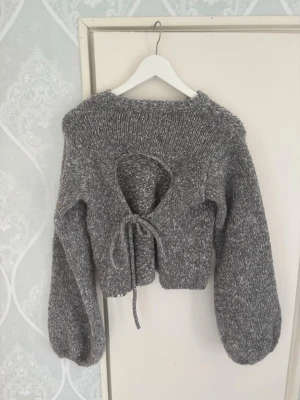 Grå stickad tröja med öppen rygg - Trendig gråmelerad stickad tröja med ballongärmar och öppen rygg som knyts med ett band. Kortare modell med bred ribbstickad kant vid halsen. Perfekt för dig som vill ha en unik detalj på din outfit.