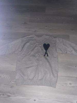 Grå stickad tröja med hjärtdetalj - Snygg grå stickad tröja med svart hjärta och bokstaven A på bröstet. Tröjan har rund halsringning, långa ärmar och ribbade muddar vid ärmslut och nederkant. Perfekt för dig som gillar enkel men trendig design.