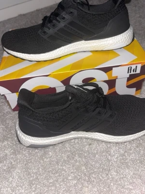 Svarta Adidas Ultra Boost sneakers - Säljer ett par svarta Adidas Ultra Boost sneakers med stickad ovandel och klassiska tre ränder på sidan. Skorna har en vit, dämpad Boost-sula och svarta detaljer i plast runt hälen och snörningen. Perfekta för dig som gillar stilrena och bekväma sneakers.