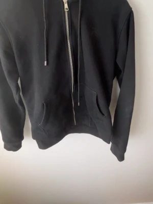Svart hoodie med dragkedja och huva - Säljer en svart hoodie från Burberry med dragkedja framtill, justerbar huva med snören och klassiskt rutigt mönster på insidan av huvan. Hoodien har känguruficka och ribbade muddar vid ärmar och nederkant. Perfekt för en chill och stilren look.