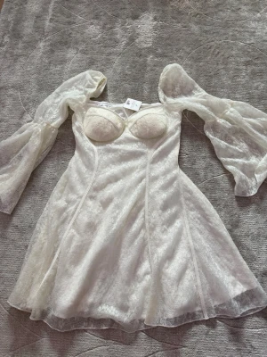 Vit kort klänning med puffärmar - Supersöt vit kort klänning med transparenta puffärmar och markerad byst. Klänningen har en lätt glansig yta och är sydd i ett tunt, skirt material som ger en romantisk känsla. Perfekt för dig som vill sticka ut med en feminin och trendig look.