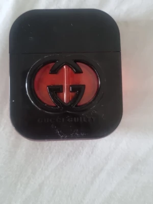 Gucci Guilty parfym svart/röd - Gucci Guilty parfym i en stilren svart flaska med det ikoniska GG-emblemet i mitten. Flaskan har en fyrkantig form med rundade hörn och ett fönster i rött glas som visar parfymens innehåll. Perfekt för dig som gillar exklusiva dofter och snygg design.