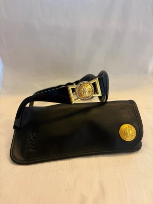 Vintage Versace solglasögon med gulddetalj - Coola svarta solglasögon från Versace med ovala glas och en ikonisk guldfärgad Medusa-detalj på sidorna. Bågarna är i blank svart plast och kommer med ett svart fodral med guldfärgad logga. Perfekt accessoar för att lyfta din stil.             Alla diamanter finns kvar .