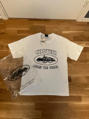 Vit Corteiz t-shirt med tryck - Säljer en vit t-shirt från Corteiz med svart tryck på bröstet där det står 'Corteiz Rules The World' och en illustration av en byggnad. T-shirten har rund hals och korta ärmar. Perfekt för dig som gillar streetwear och vill sticka ut. står M fast passar för S