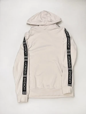 Vit hoodie från Givenchy med logoband - Snygg vit hoodie från Givenchy med svarta logoband längs ärmarna där det står GIVENCHY i vitt. Hoodien har huva med snörning och en stor magficka framtill. Perfekt för dig som vill ha en clean men ändå lyxig streetwear-look.