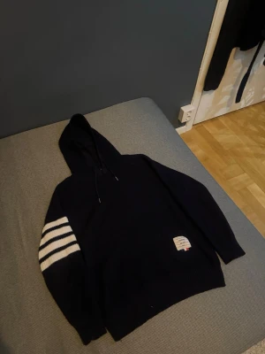 Stickad Mörkblå hoodie från Thom Browne - Snygg stickad mörkblå hoodie från Thom Browne. Ena ärmen har tre vita ränder som ger en sportig vibe. Liten patch på magen för extra detalj. Perfekt för dig som gillar en clean men ändå unik stil.