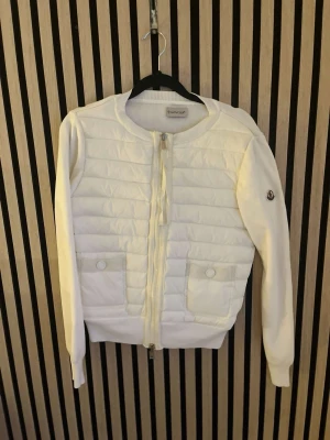 Vit quiltad cardigan från Moncler - Snygg vit cardiganjacka från Moncler med quiltat framstycke, ribbade muddar och två fickor med knapp framtill. Dragkedja hela vägen och rund halsringning. Moncler-logga på ärmen. Perfekt för lager på lager och stilren look.