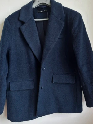 Mörkblå kavajkappa från Gina Tricot – Ullmix-look - Säljer en jättefin, mörkblå (marinblå) kavajkappa från Gina Tricot  Märke: Gina Tricot  Storlek S  Jackan har en grov, texturerad struktur (ullmix-känsla), klassiska slag, två knappar framtill och dekorativa ficklock  Skick: Använd men fint skick  Material: Texturerat material med innerfoder  Djur- och rökfritt hem. Skickas snabbt!