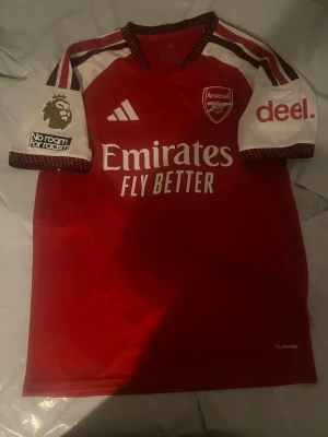 Arsenal matchtröja Saka #7 röd/vit - Snygg Arsenal matchtröja i rött och vitt med Saka #7 på ryggen. Tröjan har klubbmärke, Adidas-logga och sponsortryck på bröstet samt Premier League-märke på ärmarna. Tillverkad i lätt och ventilerande material, perfekt för fotbollsträning eller match.