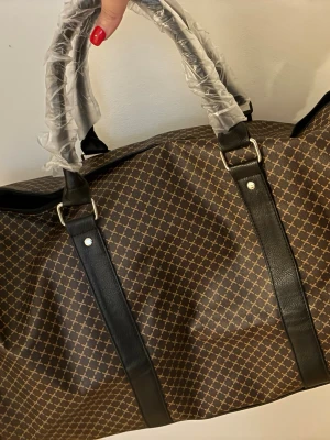 Brun weekendbag med mönster - Snygg brun weekendbag med beige mönster och svarta detaljer. Väskan har två handtag och dragkedja upptill. Materialet ser ut att vara konstläder och textil, perfekt för resor när du vill ha stil och funktion. Rymmer mycket och har klassisk design.