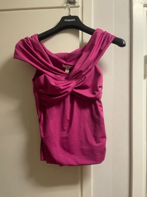 Rosa omlottopp med twistad detalj - Säljer en snygg rosa topp med omlott och twistad detalj framtill. Toppen har breda axelband och en draperad look som ger en unik stil. Perfekt för dig som vill sticka ut med färg och form.