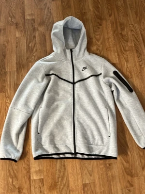 Grå Nike tracksuit med huva - Säljer ett grått tracksuit-set från Nike med svart logga. Hoodien har dragkedja, huva och svarta detaljer, samt fickor och en armficka med dragkedja. Byxorna har dragsko i midjan, muddar vid benslut och svarta detaljer på sidan. Perfekt för träning eller chill.