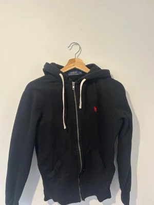 Svart hoodie från Polo Ralph Lauren - Svart hoodie från Polo Ralph Lauren med dragkedja, vit dragsko i huvan och den klassiska röda loggan broderad på bröstet. Hoodien har fickor framtill och är tillverkad i mjukt material som passar perfekt för en chill look.