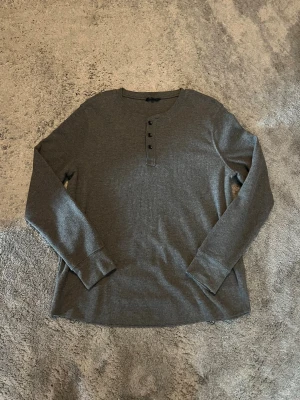 Mörkgrå långärmad henleytröja - Säljer en mörkgrå långärmad henleytröja med rund halsringning och tre knappar framtill. Tröjan är ribbad och har en avslappnad passform, perfekt för lager på lager. Materialet känns mjukt och stretchigt och denna är iprincip oanvänd! Skriv om du har frågor😇