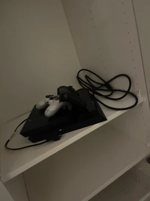 Playstation 4 med två handkontroller - Säljer en svart Playstation 4 med två trådlösa handkontroller, en vit och en svart. Konsolen har en stilren, rektangulär design och medföljer gör strömkabel och HDMI-kabel. Perfekt för gamingkvällar och multiplayer.