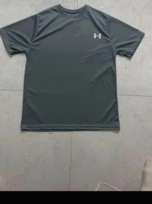 Mörkgrå tränings-tshirt Under Armour - Snygg mörkgrå tränings-tshirt från Under Armour med klassisk logga på bröstet. Tillverkad i ett lätt och snabbtorkande funktionsmaterial som andas, perfekt för gymmet eller löprundan. Rund halsringning och korta ärmar.