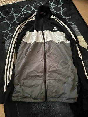 Svart och grå Adidas träningsjacka - Snygg träningsjacka från Adidas i svart, grått och vitt. Jackan har dragkedja framtill, ståkrage och klassiska vita ränder längs ärmarna. Tillverkad i Primegreen-material och har Adidas-logga på bröstet. Perfekt för träning eller chill.