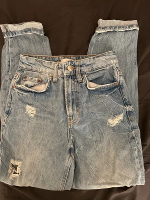 Ljusblå slitna mom jeans - Säljer ett par ljusblå mom jeans med hög midja och flera slitningar och hål på både fram- och baksida. Jeansen har fem fickor, knappgylf och uppvikta benslut. Perfekta för en avslappnad och trendig look. Är i storlek 6.