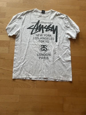 Vit Stüssy t-shirt med tryck - Vit t-shirt från Stüssy med svart tryck fram och bak. Framsidan har Stüssy-loggan och namn på storstäder, medan baksidan har logga och fler stadsnamn. Klassisk passform och rund hals. Perfekt för dig som gillar streetwear.