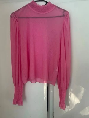Rosa transparent blus med puffärm - Säljer en rosa transparent blus med hög krage och långa puffärmar. Blusen har smockdetaljer vid halsen och ärmsluten, vilket ger en trendig och luftig look. Perfekt för dig som gillar färg och vill sticka ut lite extra.