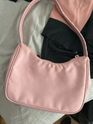 Ljusrosa axelremsväska - Säljer en ljusrosa handväska i mjukt, slätt material med enkel design. Väskan har en avlång form och en bred axelrem. Stängs med dragkedja upptill och har en stilren look som passar till många outfits.