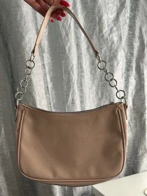 Beige axelremsväska med metallringar - Stilren beige handväska i slätt, glansigt material med axelrem dekorerad med stora silverfärgade metallringar. Väskan har en mjukt rundad form och dragkedja upptill. Perfekt för dig som gillar minimalistisk och trendig design.