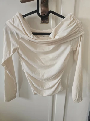 Vit offshoulder topp från H&M - Säljer en vit offshoulder topp från H&M i mjukt material. Toppen har långa ärmar och en snygg draperad ringning som ger en trendig look. Perfekt att styla med jeans eller kjol för en cool outfit.