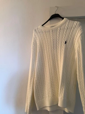 Polo sweatshirt  - Kabelstickad vit tröja med rund hals och långa ärmar. Tröjan har ett broderat logotypmärke på bröstet och ribbstickade muddar vid ärmslut och nederkant. Perfekt för en stilren och klassisk look.